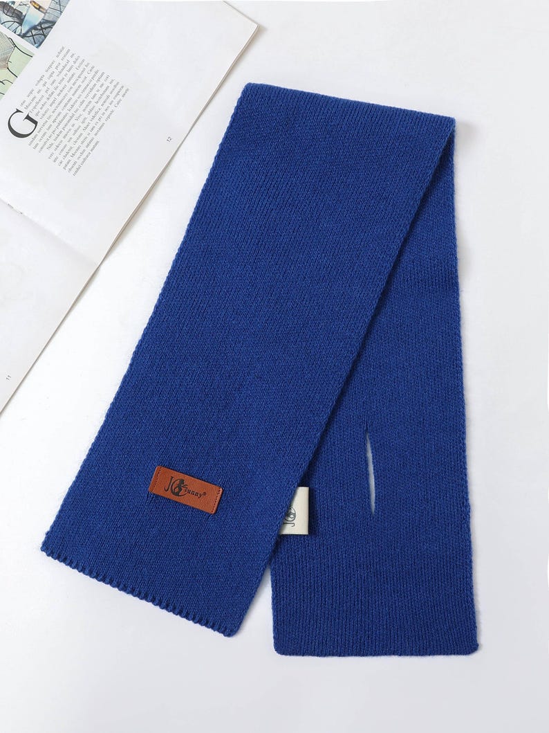 Puede incluir: Una bufanda de punto azul real con forma rectangular y una abertura de paso. Una etiqueta de cuero marr&oacute;n con el texto "JG Supply" est&aacute; adjunta. La bufanda est&aacute; doblada sobre una superficie blanca.