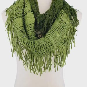 May include: A green infinity scarf with a fringed edge. The scarf is made of a knitted material and is displayed on a mannequin. The scarf has a textured appearance and is a solid green color.