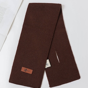 Puede incluir: Bufanda de punto marr&oacute;n oscuro con una etiqueta rectangular de cuero que dice "JG Jenny". La bufanda est&aacute; doblada por la mitad, con una peque&ntilde;a abertura. Una etiqueta blanca est&aacute; adjunta.