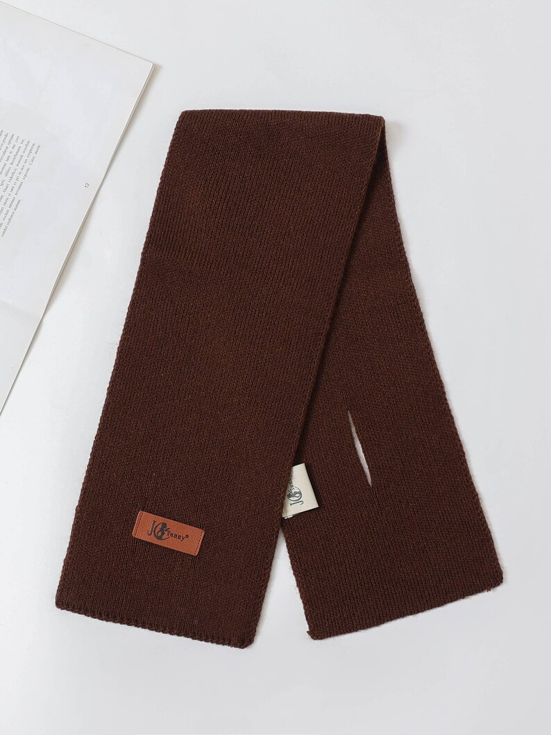 Puede incluir: Bufanda de punto marr&oacute;n oscuro con una etiqueta rectangular de cuero que dice "JG Jenny". La bufanda est&aacute; doblada por la mitad, con una peque&ntilde;a abertura. Una etiqueta blanca est&aacute; adjunta.