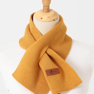 Puede incluir: Bufanda de punto amarillo mostaza exhibida en un maniqu&iacute;. La bufanda est&aacute; envuelta alrededor del cuello y tiene un parche rectangular con el texto "JG Sunny". La bufanda est&aacute; hecha de un material suave y texturizado.