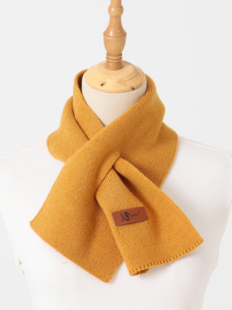 Puede incluir: Bufanda de punto amarillo mostaza exhibida en un maniqu&iacute;. La bufanda est&aacute; envuelta alrededor del cuello y tiene un parche rectangular con el texto "JG Sunny". La bufanda est&aacute; hecha de un material suave y texturizado.