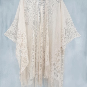 May include: Off-white lace kimono with floral patterns and fringe trim. The open-front design features wide sleeves and a delicate, airy texture. A stylish, bohemian-inspired garment.