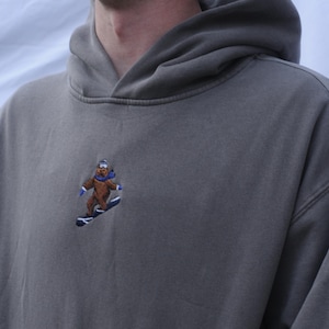 Puede incluir: Una sudadera con capucha gris con un oso marrón que lleva un gorro azul y practica snowboard en una tabla de snowboard azul bordada en la parte delantera.