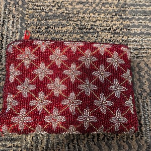 May include: A red beaded clutch with a gold zipper and a floral pattern. The clutch is made of small, round beads that are sewn together to create a textured surface.