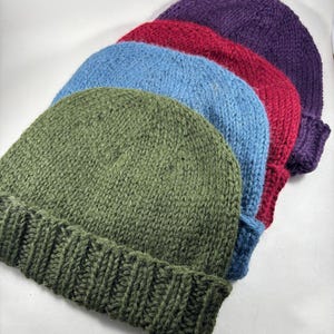 Puede incluir: Un conjunto de cuatro gorros de punto apilados en fila, con un borde acanalado y una corona de punto liso. Los gorros son de color verde oliva, azul claro, rojo oscuro y morado oscuro, cada uno elaborado con una textura de lana gruesa.