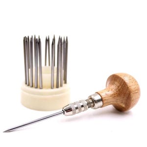 May include: A set of jewelry making tools. A wooden handled awl with a silver tip and a set of silver needles in a cream-colored holder are displayed against a white background. The awl is in focus, with the needles slightly blurred.