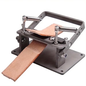 May include: A metal leather skiving machine with a rectangular base and a square frame. Two springs are attached to the frame. A piece of brown leather is placed in the machine, with a second piece of leather extending out.