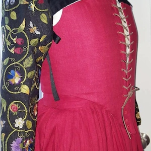 May include: A red linen dress with a black velvet bodice and embroidered sleeves. The bodice has a lace-up back and the sleeves feature floral and bee embroidery.