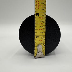 May include: A black round object being measured with a yellow tape measure. The tape measure reads 1 3/4 inches.