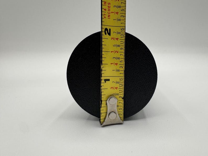 May include: A black round object being measured with a yellow tape measure. The tape measure reads 1 3/4 inches.