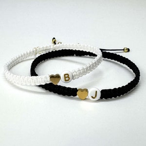May include: Two friendship bracelets, one white and one black, with gold heart charms and letter beads. The white bracelet has a "B" bead and the black bracelet has a "J" bead.