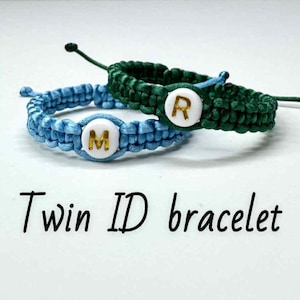 Peut inclure: Deux bracelets en macramé, un bleu et un vert, chacun avec un charme circulaire blanc avec une lettre dorée. Le bracelet bleu porte la lettre "M", et le bracelet vert porte la lettre "R". Le texte "Twin ID bracelet" est en dessous.