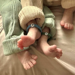 May include: Close-up of two pairs of baby feet with blue and green bracelets. The bracelets have white letter beads with the letters "P" and "L". The babies are wearing light green and beige clothing.