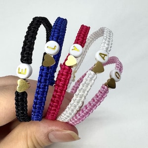 Peut inclure: Cinq bracelets d'amitié colorés avec des perles blanches portant des lettres dorées. Chaque bracelet est orné d'un charm en forme de cœur doré.