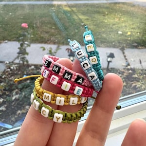 May include: A stack of five woven cord bracelets in teal, pink, yellow, and olive green. Each bracelet features square white beads with black or gold letters spelling out the names EMMA, SOFIE, MILA, LUCY, and LUCA, with a teal bracelet on top.