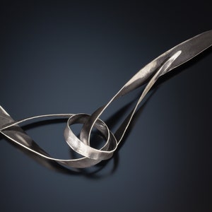 May include: A silver necklace featuring a distinctive, abstract design. The necklace is crafted from a single piece of metal that is twisted and turned into an intricate shape.