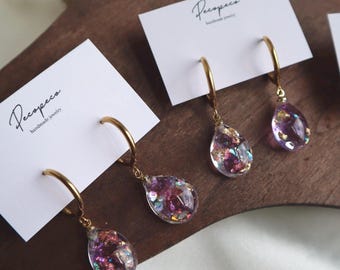 May include: Gold hoop earrings with teardrop-shaped pendants. The clear pendants are filled with colorful glitter and iridescent flakes. The earrings are displayed on white cards with the text "Pecopeco handmade jewelry".