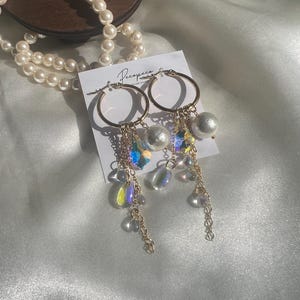 Puede incluir: Pendientes de aro dorados con cristales iridiscentes, perlas y delicadas cadenas. Los pendientes se exhiben en una tarjeta blanca con el nombre de la marca "Recopaco" en escritura cursiva. Un collar de perlas es visible en el fondo.