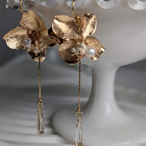May include: A pair of gold-colored floral earrings. Each earring features a detailed flower design with pearl accents and a dangling clear crystal teardrop. The earrings are displayed against a white background.