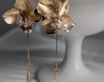 May include: A pair of gold-colored floral earrings. Each earring features a detailed flower design with pearl accents and a dangling clear crystal teardrop. The earrings are displayed against a white background.