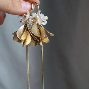 May include: Gold-toned earrings featuring a floral design. The earrings have small white flower clusters at the top, with textured gold leaf-shaped dangles and long, delicate gold chains. The earrings are designed to dangle.