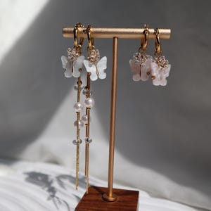 May include: Gold hoop earrings with butterfly charms. One pair features white butterfly pendants with pearl and crystal accents, and another pair has pink butterfly pendants. The earrings are displayed on a gold stand with a wooden base.
