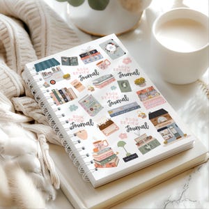 May include: A white spiral-bound notebook with a cover featuring colorful illustrations of books, mugs, and the text "Book Review Journal." The notebook is open on top of a closed book, with a mug of a milky beverage in the background.