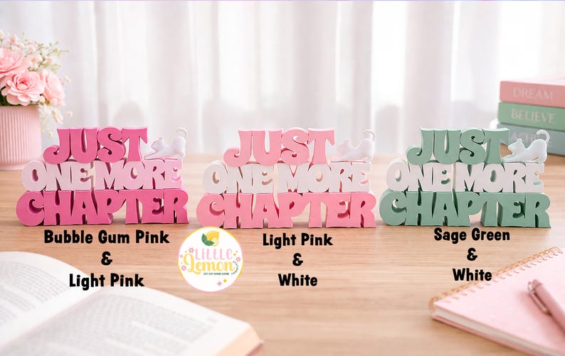 May include: Three decorative wooden signs with the text "JUST ONE MORE CHAPTER" in bubble gum pink, light pink, and sage green. Each sign has a small white cat figurine on top. A book and notepad are also visible.