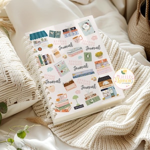 May include: A spiral-bound journal with a white cover featuring colorful illustrations of books, mugs, and decorative elements. The words "Reading Tracker Journal" are repeated throughout the design. The journal is open, displaying the pages, and rests on a cream-colored knitted blanket.