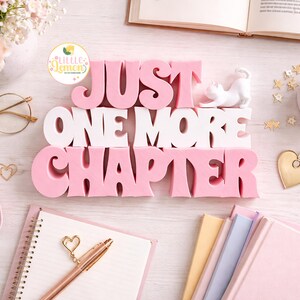 May include: A flat lay featuring a pink and white decorative sign that reads "JUST ONE MORE CHAPTER." The scene includes a cup of coffee, books, glasses, and stationery, all in soft, pastel colors. Gold accents add a touch of elegance.