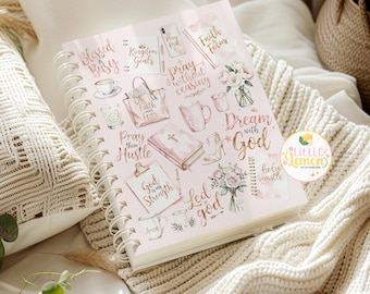 May include: A spiral-bound journal with a light pink cover featuring various illustrations and text in gold and pink. The journal includes images of a coffee cup, handbag, bible, and flowers. Text includes "Blessed", "Dream with God", and "Pray without ceasing".
