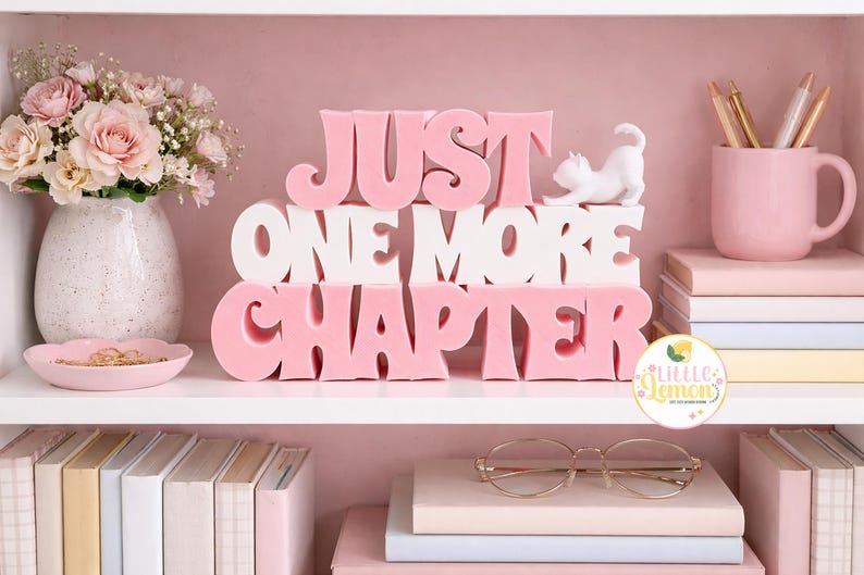 May include: A pink and white decorative shelf arrangement with the words "JUST ONE MORE CHAPTER". The shelf also features a vase of pink flowers, a pink mug with pens, books, and a small white cat figurine.