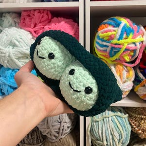 Puede incluir: Una vaina de guisantes de crochet verde con dos guisantes verdes sonrientes dentro. Los guisantes tienen ojos negros y una sonrisa cosida. La vaina se sostiene en una mano.