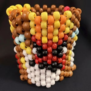 May include: A handmade beaded cuff bracelet featuring a colorful design. The bracelet is primarily brown, yellow, orange, red, black, and white. The beads are arranged to create a face-like pattern. The cuff is cylindrical and appears to be made of plastic beads.