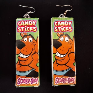 Puede incluir: Un par de pendientes originales hechos con cajas de Candy Sticks recicladas. Cada pendiente presenta una imagen de dibujos animados de Scooby-Doo sobre un fondo verde, con las palabras "CANDY STICKS" y "SCOOBY-DOO!" impresas. Ganchos para las orejas plateados.