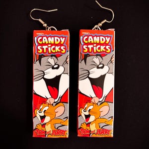 May include: A pair of novelty earrings featuring the iconic Tom and Jerry cartoon characters. The earrings are rectangular, with a red background and the words "CANDY STICKS" in yellow and blue. The design includes images of Tom the cat and Jerry the mouse.