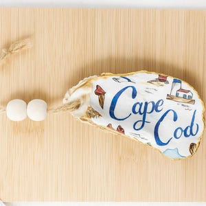 以下が含まれることがあります： ケープコッドのデザインが施されたカキの殻。青い文字とランドマークのイラストが描かれています。2つの白い木製ビーズが付いた天然の紐が付いています。殻は、明るい茶色の木製まな板の上に置かれています。