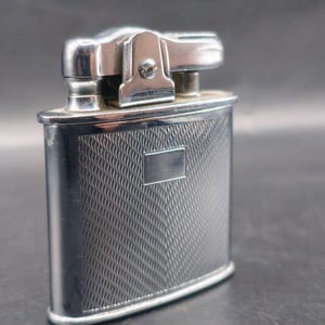 May include: A vintage silver-toned lighter with a rectangular body and a textured, patterned design. The top features a hinged mechanism and a small screw. The lighter has a rectangular plate on the front.