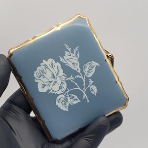 May include: A square compact case featuring a matte dusty blue surface with a white floral rose illustration in the center. The compact is framed by a scalloped gold-tone metal edge with a hinge mechanism visible on the right side.