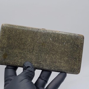 Op de afbeelding: Een platte, rechthoekige etui met een getextureerd, olijfgroen oppervlak met een fijn, honingraatachtig patroon. De randen van de etui zijn afgewerkt met een metalen bies, wat een slank profiel en een scharnierconstructie voor het openen toont.