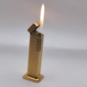 May include: A gold-coloured, rectangular lighter with a textured surface. The lighter is open, with a flame burning upwards. The base of the lighter is square, and the top is rectangular. The lighter is set against a white background.