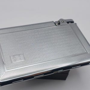 May include: A rectangular, silver cigarette case with a textured pattern and a hinged lid. The case has a small, rectangular area with the initials "K G" and "W G" engraved. The case is made of a shiny, metallic material.
