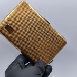 Puede incluir: Un pitillera rectangular de color dorado con un patrón texturizado de trama cruzada. La pitillera tiene un pequeño acento rectangular en la parte delantera y un cierre de broche. La pitillera está sujeta por una mano enguantada.