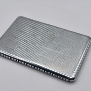May include: A rectangular, silver-colored cigarette case with a textured, striped pattern. The case has a small, rectangular area on the front with a smooth finish. The edges are rounded and the case appears to be made of metal.
