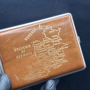 May include: A vintage brown leather cigarette case with a silver metal frame. The case is embossed with a map of Northern Germany and the words "Souvenir of Germany" and "Nordsee".