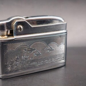 May include: A rectangular silver-tone metal lighter featuring an engraved scene on its front surface. The engraving depicts a landscape with birds flying over a body of water and stylized clouds above. The lighter has a smooth, polished finish.