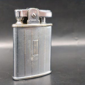 May include: A vintage silver-toned lighter with a rectangular body and a hinged top. The lighter features vertical grooved detailing on the front and a rectangular accent. The word "Ronson" is visible on the top.