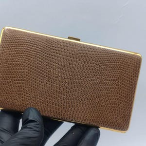 Pode incluir: Uma bolsa clutch retangular com exterior castanho e um padrão texturizado de pele de lagarto. A bolsa é emoldurada por uma borda de metal dourado polido e inclui um pequeno fecho retangular de metal dourado na borda superior.