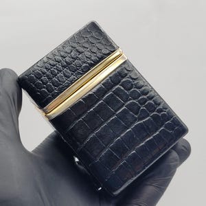以下が含まれることがあります： 黒い外装にクロコダイル柄の型押しが施された長方形のシガレットケース。中央の開口部に沿って細い光沢のあるゴールドトーンの金属トリムがあしらわれており、暗いエンボス加工の表面と対照的なアクセントになっています。